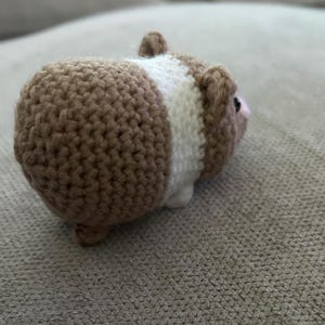 Op de afbeelding: Een klein, gehaakt knuffeldier in de vorm van een cavia. Het speelgoed heeft een rond lichaam met een textuur van beige garen, een brede witte band rond het midden en kleine beige oortjes. Het heeft een zichtbaar donker oog en een roze neus.