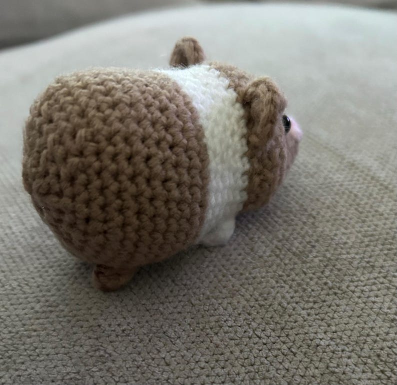 Op de afbeelding: Een klein, gehaakt knuffeldier in de vorm van een cavia. Het speelgoed heeft een rond lichaam met een textuur van beige garen, een brede witte band rond het midden en kleine beige oortjes. Het heeft een zichtbaar donker oog en een roze neus.