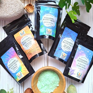 May include: A selection of aromatherapy foaming bath salts in black pouches, each labelled with different scents such as "Muscle Ease" and "Calm the Fork Down." The pouches are 70g each. A wooden bowl contains green bath salts.