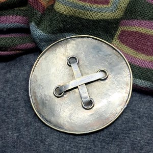 Może przedstawiać: Duży, okrągły metalowy guzik z młotkowanym, srebrzystym wykończeniem. Guzik posiada centralny detal w kształcie krzyża, utworzony przez dwie przecinające się metalowe listwy, które łączą się z czterema okrągłymi oczkami na powierzchni.