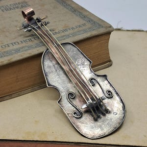 May include: A silver and copper double bass pendant with intricate details. The pendant is shaped like a double bass and has a textured surface. The strings are made of copper wire.
