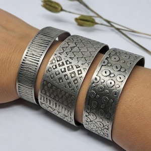 Pode incluir: Três pulseiras de metal em tom prateado com padrões gravados distintos. Uma apresenta linhas verticais, outra mostra uma mistura de triângulos, losangos e pontos, e a terceira é coberta por um padrão repetido de pequenos círculos irregulares.