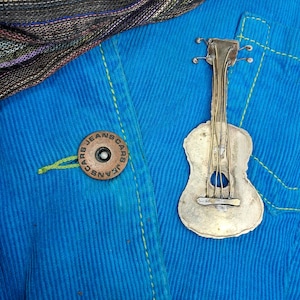 以下が含まれることがあります： 青いデニムの背景に、銀色の金属製のギターのピンが置かれています。ピンは、緑色の糸の縫い目のある青いコーデュロイのジャケットに取り付けられています。ジャケットには、「Jeans Cars」という文字が書かれたボタンが付いています。