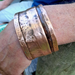 Pode incluir: Uma pulseira larga de cobre martelado com um acabamento rústico e texturizado. A superfície metálica apresenta variações de cor, variando de tons de cobre metálico brilhante a manchas mais escuras e oxidadas por toda a pulseira.
