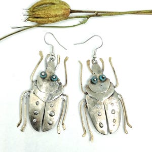 May include: A pair of silver-toned metal beetle earrings featuring a textured finish, thin wire legs, and small turquoise-colored circular accents for eyes. The earrings hang from standard silver-toned metal ear wires.