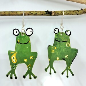 Pode incluir: Brincos de sapo feitos à mão. Cada brinco apresenta um sapo verde com manchas amarelas, olhos pretos e uma boca sorridente. Os brincos são suspensos por ganchos de orelha prateados e estão pendurados num galho castanho.
