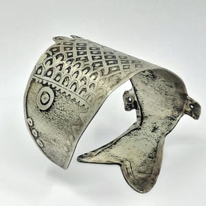 Könnte beinhalten: Ein silberfarbenes Armband in Fischform mit oxidiertem Finish und eingravierten Mustern, darunter ein gitterartiges Netz, schuppenartige Motive und kreisförmige Details auf dem Körper, das in einer schwanzförmigen Öffnung endet.