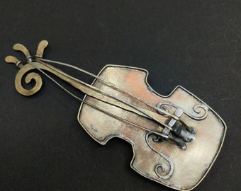 Op de afbeelding: Een metalen broche in de vorm van een viool met een bruine en zilveren afwerking. De viool heeft een gedetailleerd ontwerp met ingewikkelde swirls en lijnen.