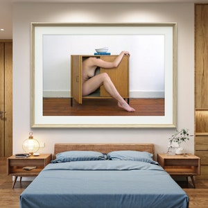 Puede incluir: Una obra de arte enmarcada representa a una persona dentro de un armario de madera con las puertas abiertas. El armario est&aacute; contra una pared blanca, con un cuenco y libros encima. La obra cuelga sobre una cama con ropa de cama azul y mesitas de noche de madera.