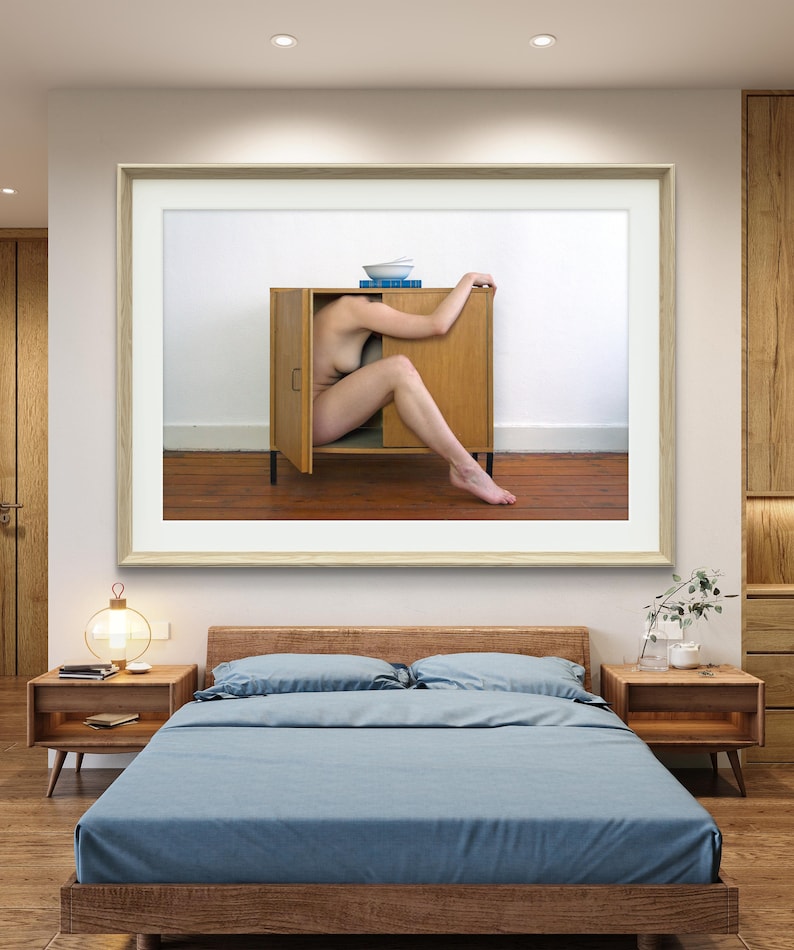 Puede incluir: Una obra de arte enmarcada representa a una persona dentro de un armario de madera con las puertas abiertas. El armario est&aacute; contra una pared blanca, con un cuenco y libros encima. La obra cuelga sobre una cama con ropa de cama azul y mesitas de noche de madera.
