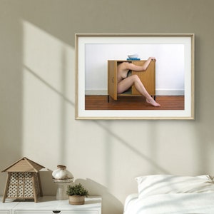 Puede incluir: Fotograf&iacute;a enmarcada de una persona dentro de un armario de madera abierto. Un cuenco azul y un libro est&aacute;n encima. La foto est&aacute; colgada en una pared beige. Una mesita de noche blanca, una l&aacute;mpara decorativa y una cama con edred&oacute;n blanco est&aacute;n en primer plano.