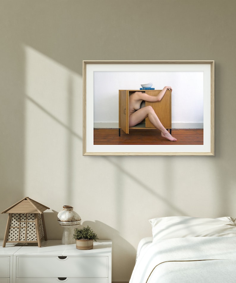 Puede incluir: Fotograf&iacute;a enmarcada de una persona dentro de un armario de madera abierto. Un cuenco azul y un libro est&aacute;n encima. La foto est&aacute; colgada en una pared beige. Una mesita de noche blanca, una l&aacute;mpara decorativa y una cama con edred&oacute;n blanco est&aacute;n en primer plano.