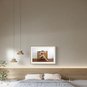 Puede incluir: Fotograf&iacute;a enmarcada que muestra a una persona parcialmente dentro de un armario de madera. La puerta del armario est&aacute; abierta, revelando las piernas y el torso de la persona. La foto cuelga en una pared neutra sobre una cama con una colcha a rayas.