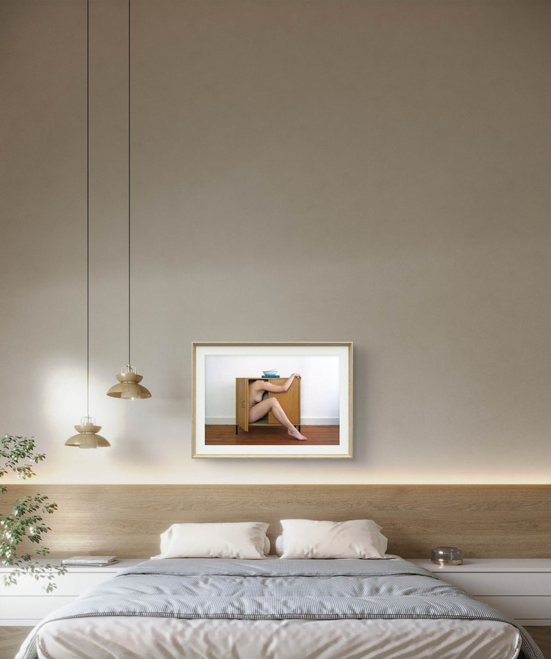 Puede incluir: Fotograf&iacute;a enmarcada que muestra a una persona parcialmente dentro de un armario de madera. La puerta del armario est&aacute; abierta, revelando las piernas y el torso de la persona. La foto cuelga en una pared neutra sobre una cama con una colcha a rayas.