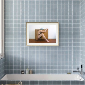 Puede incluir: Una fotograf&iacute;a enmarcada cuelga de una pared de azulejos azules en un ba&ntilde;o. La fotograf&iacute;a representa a una persona dentro de un armario de madera. El ba&ntilde;o cuenta con una ba&ntilde;era blanca, una ventana y una cesta.