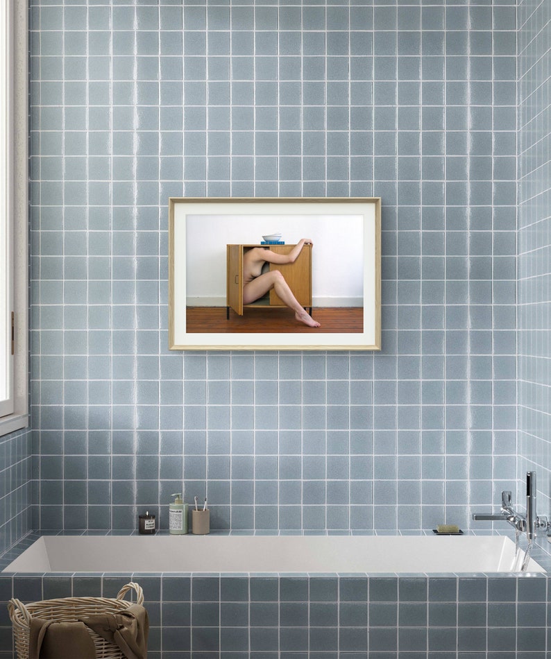 Puede incluir: Una fotograf&iacute;a enmarcada cuelga de una pared de azulejos azules en un ba&ntilde;o. La fotograf&iacute;a representa a una persona dentro de un armario de madera. El ba&ntilde;o cuenta con una ba&ntilde;era blanca, una ventana y una cesta.