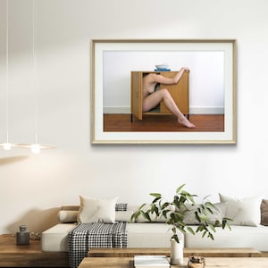 Puede incluir: Impresi&oacute;n art&iacute;stica enmarcada que muestra a una persona dentro de un armario de madera con una puerta abierta. El armario est&aacute; contra una pared blanca y un suelo marr&oacute;n. Un cuenco azul y un libro encima.