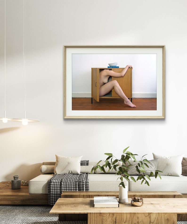 Puede incluir: Impresi&oacute;n art&iacute;stica enmarcada que muestra a una persona dentro de un armario de madera con una puerta abierta. El armario est&aacute; contra una pared blanca y un suelo marr&oacute;n. Un cuenco azul y un libro encima.