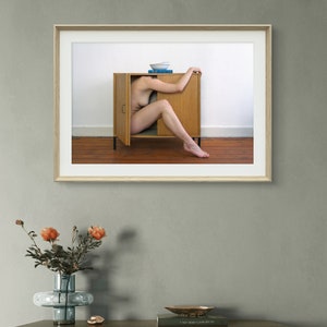 Puede incluir: Una impresi&oacute;n art&iacute;stica enmarcada que muestra a una persona dentro de un armario de madera abierto. Un cuenco blanco y libros azules est&aacute;n encima. La obra de arte cuelga en una pared gris sobre un mueble de madera. Un jarr&oacute;n con flores naranjas est&aacute; en el mueble.
