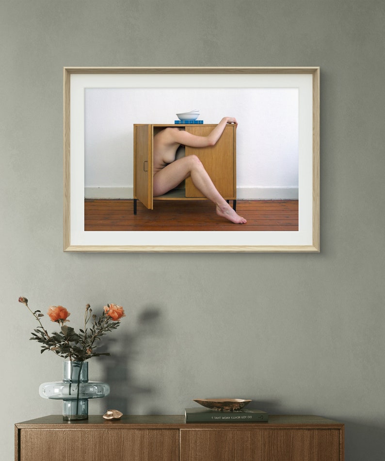 Puede incluir: Una impresi&oacute;n art&iacute;stica enmarcada que muestra a una persona dentro de un armario de madera abierto. Un cuenco blanco y libros azules est&aacute;n encima. La obra de arte cuelga en una pared gris sobre un mueble de madera. Un jarr&oacute;n con flores naranjas est&aacute; en el mueble.