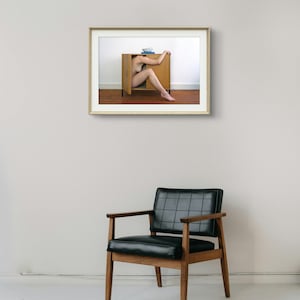 Puede incluir: Una fotograf&iacute;a enmarcada muestra a una persona dentro de un armario de madera, con un cuenco azul y libros encima. El armario est&aacute; contra una pared blanca y un suelo marr&oacute;n. Una silla de cuero negro con brazos y patas de madera est&aacute; en primer plano.