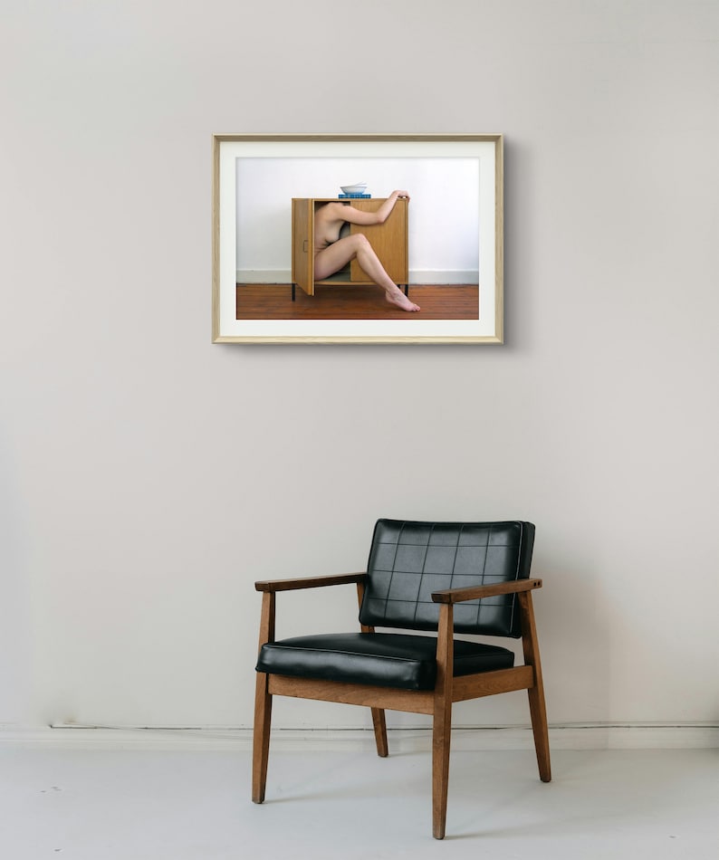 Puede incluir: Una fotograf&iacute;a enmarcada muestra a una persona dentro de un armario de madera, con un cuenco azul y libros encima. El armario est&aacute; contra una pared blanca y un suelo marr&oacute;n. Una silla de cuero negro con brazos y patas de madera est&aacute; en primer plano.