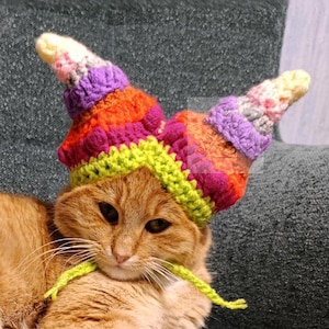 May include: An orange tabby cat wearing a colorful, crocheted hat with two cone-shaped points. The cat is lying on a gray sofa next to a yellow ball. The word "PATTERN" is written vertically on the left.