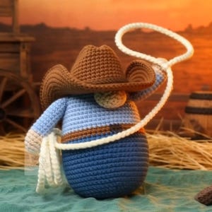 Puede incluir: Gnomo vaquero de ganchillo con sombrero marrón y camisa azul, sosteniendo un lazo de cuerda blanco. El gnomo tiene una cara bronceada y un cinturón marrón. El fondo presenta una escena de puesta de sol.