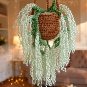 May include: A handmade crochet hanging planter with a brown pot, green vines, and trailing light green yarn. The planter is suspended by a brown rope, creating a whimsical, decorative piece for indoor plants. The overall design is a charming, handcrafted item.