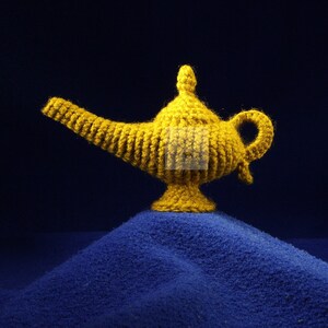 K&ouml;nnte beinhalten: Eine handgefertigte, goldgelbe H&auml;kellampe, die an eine Wunderlampe erinnert, steht auf einer blauen, strukturierten Oberfl&auml;che. Die Lampe hat einen gebogenen Ausguss, einen Griff und eine dekorative Oberseite. Das Wort "PATTERN" wird links vertikal angezeigt.