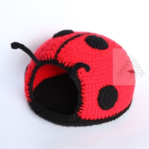 Pode incluir: Uma cama para animais de estimação em forma de joaninha, feita de crochê vermelho com manchas pretas e um interior de crochê preto. A cama tem uma pequena abertura para o animal de estimação entrar. O texto "DASHAH HOUSE" é visível no lado da cama.