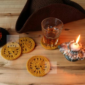 Op de afbeelding: Een stilleven met een glazen fles, een glas amberkleurige vloeistof en een bruine gehaakte hoed. Er zijn verschillende gele gehaakte onderzetters rond het glas en de fles geplaatst. Een brandende kaars staat op een decoratieve houder.