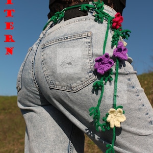 Pode incluir: Um cinto de crochê feito à mão com vinhas verdes e flores vermelhas, roxas e creme. O cinto é usado com jeans de lavagem clara. A palavra "PATTERN" é exibida verticalmente em letras vermelhas.