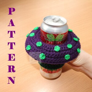 May include: A purple crocheted drink cosy with small green circular accents, featuring a wide brim that encircles a 500 ml aluminium beverage can. The word PATTERN is printed vertically in purple letters on the left side of the image.