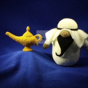 May include: A crocheted genie figure with a white robe and black beard stands next to a golden crocheted lamp. The figures are on a blue background.