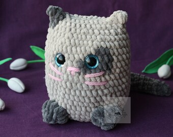 May include: A gray and black crocheted cat toy with pink yarn details for the nose and whiskers. The cat toy has blue eyes and is sitting on a purple surface with white tulips.