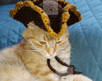 May include: A ginger cat wearing a handmade brown and gold crochet pirate hat with a matching neck tie. The cat is lying on a teal quilted blanket. The word "PATTERN" is written vertically in white letters.