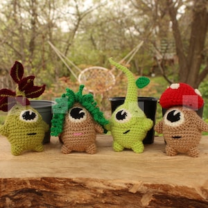 Pode incluir: Quatro figuras de crochê feitas à mão em várias cores, que se assemelham a criaturas caprichosas semelhantes a plantas. As figuras são em tons de verde, castanho e vermelho, com olhos pretos e pequenos sorrisos. Estão dispostas sobre uma superfície de madeira com um fundo exterior desfocado.