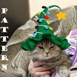 May include: A gray tabby cat wearing a green crocheted Christmas tree hat with colorful lights and a yellow star on top. The cat is sitting on a person's lap.
