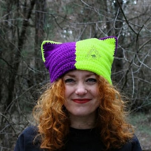May include: A handmade crochet cat ear hat in purple and neon green, worn by a person with red curly hair. The hat has a split design. The person is wearing a black top and a brown jacket. The word "PATTERN" is vertically printed on the left side.