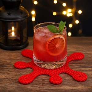 May include: A vibrant cocktail with a blood orange slice and mint garnish rests on a red crocheted coaster. A lit candle in a black lantern and fairy lights create a warm atmosphere. The drink is in a clear glass.