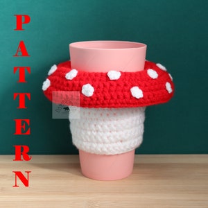 May include: A pink plastic cup featuring a crocheted mushroom cosy. The cosy has a white stem section and a red cap with white spots. The word PATTERN is written in red vertical letters on the left side of the image, and a small logo is visible.