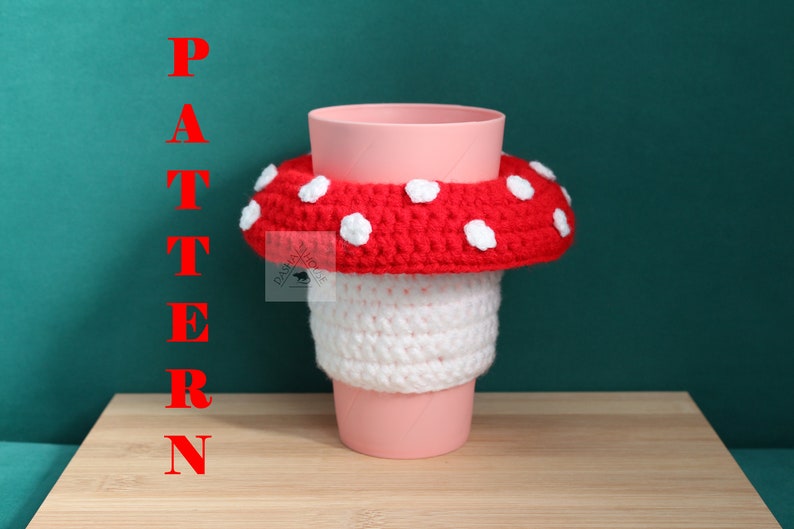 May include: A pink plastic cup featuring a crocheted mushroom cosy. The cosy has a white stem section and a red cap with white spots. The word PATTERN is written in red vertical letters on the left side of the image, and a small logo is visible.