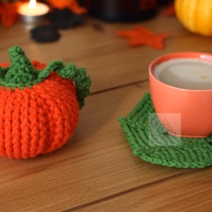 May include: An orange crocheted pumpkin-shaped item with green accents sits next to a green leaf-shaped coaster holding an orange mug filled with a beverage. The items are on a wooden surface, suggesting a fall theme.