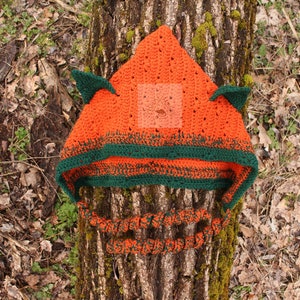 May include: A bright orange crocheted hood with two pointed ears and a dark green border. The hood features long, twisted orange and green braided ties hanging from the sides. The word PATTERN is printed vertically in white on the left side.
