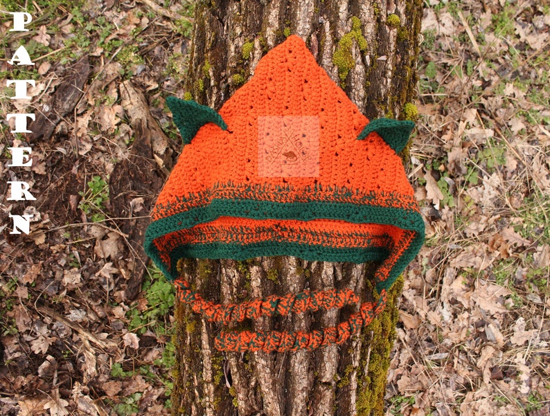 May include: A bright orange crocheted hood with two pointed ears and a dark green border. The hood features long, twisted orange and green braided ties hanging from the sides. The word PATTERN is printed vertically in white on the left side.