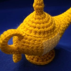 K&ouml;nnte beinhalten: Eine handgefertigte, gelbe H&auml;kellampe mit gebogenem Ausguss und rundem Sockel. Die Lampe hat einen kleinen Griff und einen wei&szlig;en Fleck an der Seite. Der Hintergrund ist einfarbig blau.