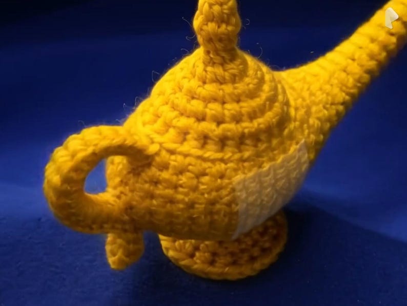 K&ouml;nnte beinhalten: Eine handgefertigte, gelbe H&auml;kellampe mit gebogenem Ausguss und rundem Sockel. Die Lampe hat einen kleinen Griff und einen wei&szlig;en Fleck an der Seite. Der Hintergrund ist einfarbig blau.