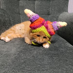 May include: An orange tabby cat wearing a colorful, crocheted hat with a spiral design. The hat features shades of yellow, green, orange, purple, and pink. The cat is lying on a gray upholstered surface.