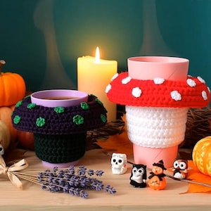 May include: Two crochet cup cozies shaped like mushrooms. One cozy is dark purple with green spots on a black base, and the other is red with white spots on a white base. They are placed around cups, surrounded by pumpkins, small figurines, and lavender.